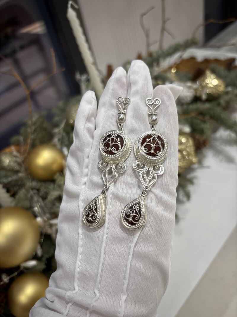 A pair of ornate silver earrings with reddish-brown stones are held in a white-gloved hand, set against a festive background with gold ornaments.