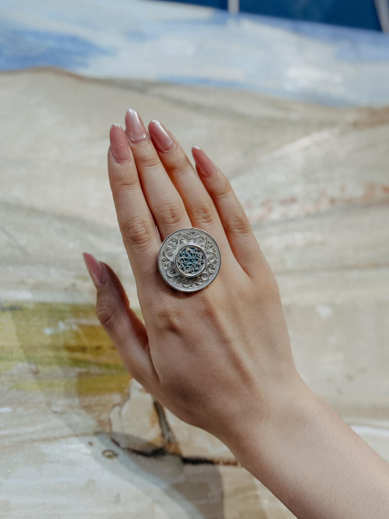 A hand with long, manicured nails is adorned with a large, round silver ring featuring intricate patterns and a turquoise-colored center.