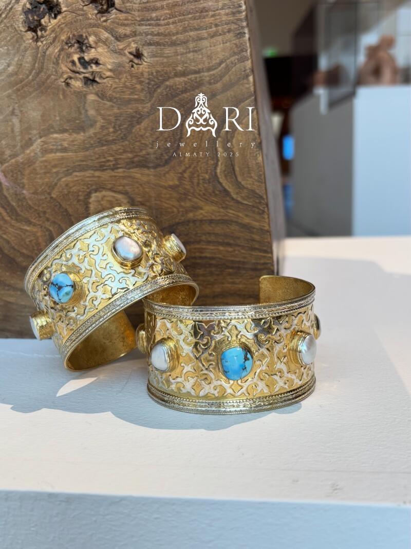Two gold bracelets decorated with turquoise and pearls are displayed next to a wooden box with the text "DARI jewellery ALMATY 2025" on it.
