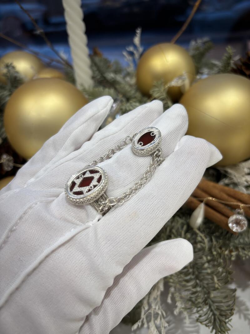 A hand in a white glove displays a silver ring connected to a silver cufflink by a chain, with a holiday-themed background featuring golden ornaments and greenery.