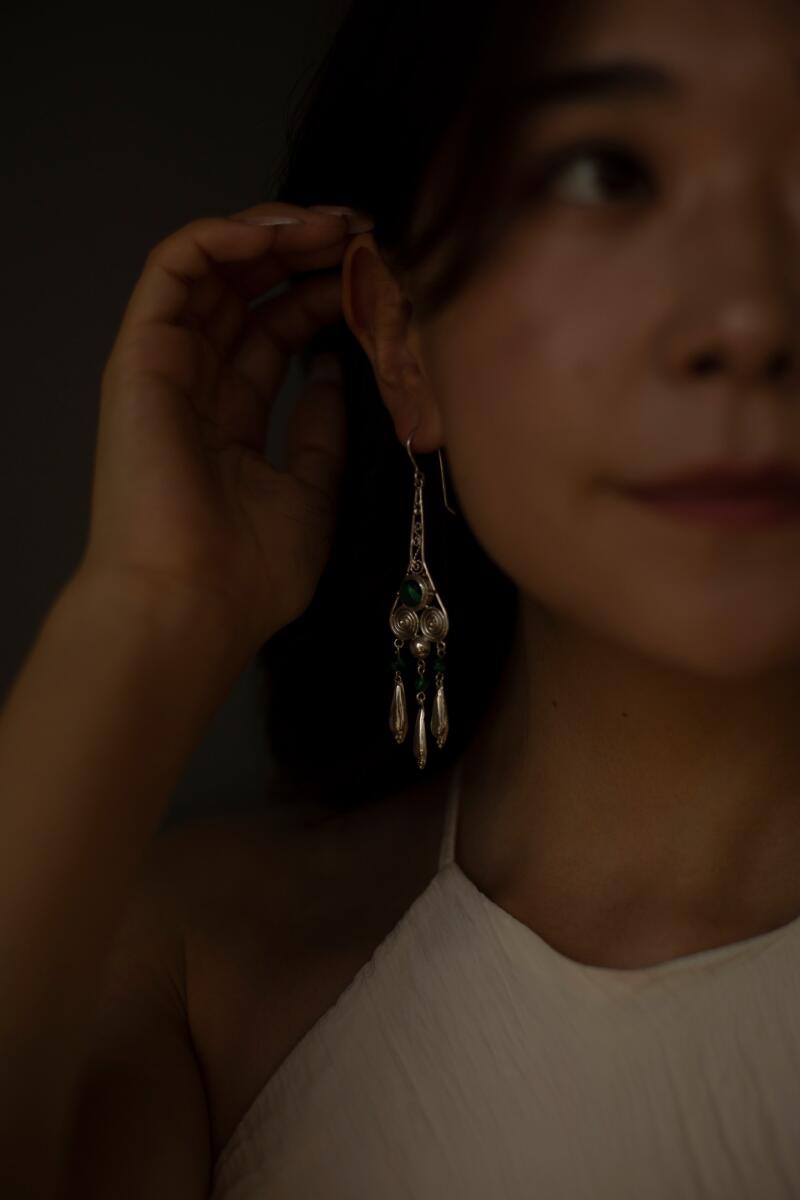 A woman with dark hair is wearing a long silver earring with green stones, touching her ear, and wearing a white halter top.