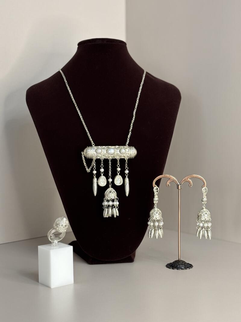 A jewelry set including a necklace, earrings, and a ring, all made of silver and pearls, are displayed against a neutral background with a dark brown velvet bust.