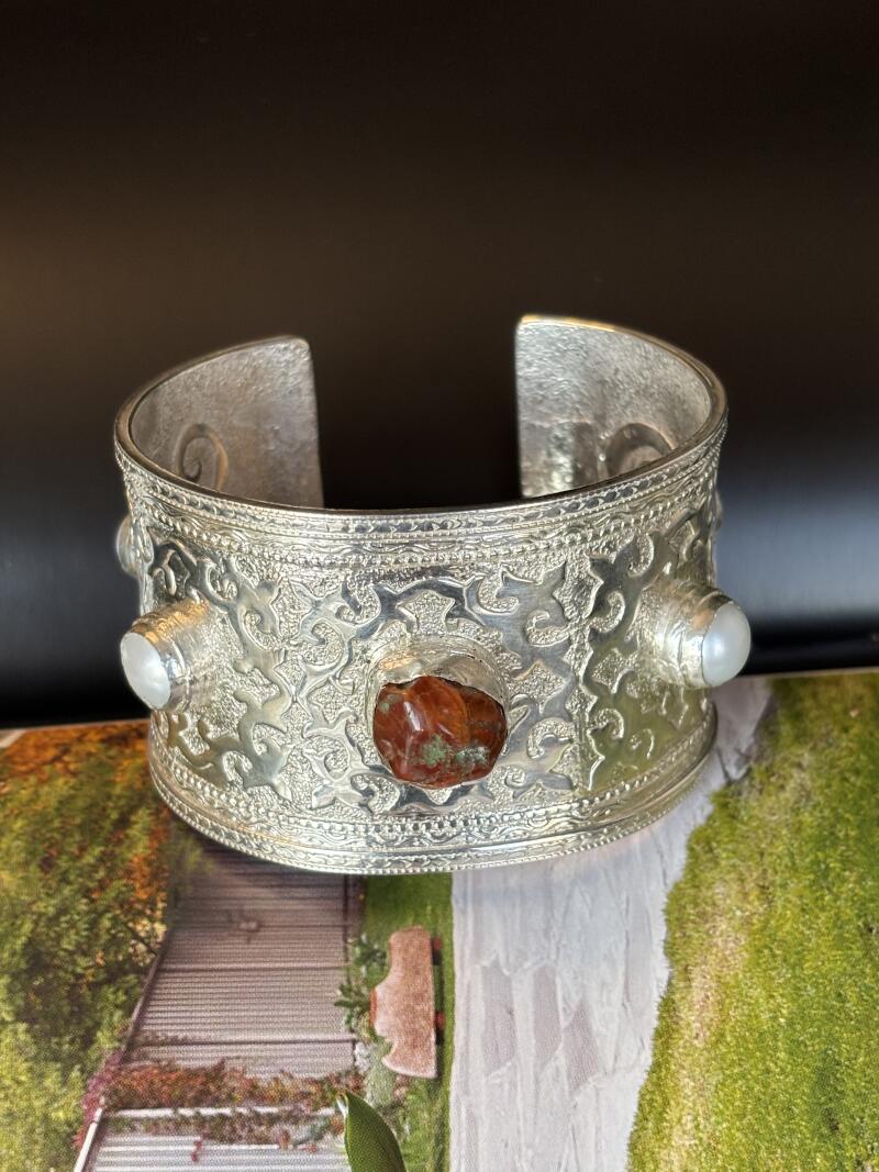 A silver cuff bracelet with a red stone in the center and white pearls on either side rests on a picture of a garden walkway.