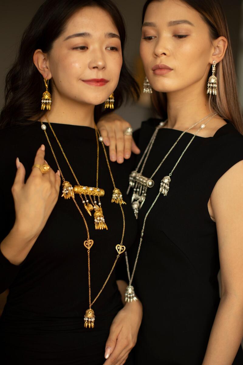 Two women are posing in black dresses and traditional Kazakh jewelry, including earrings and necklaces in gold and silver tones.