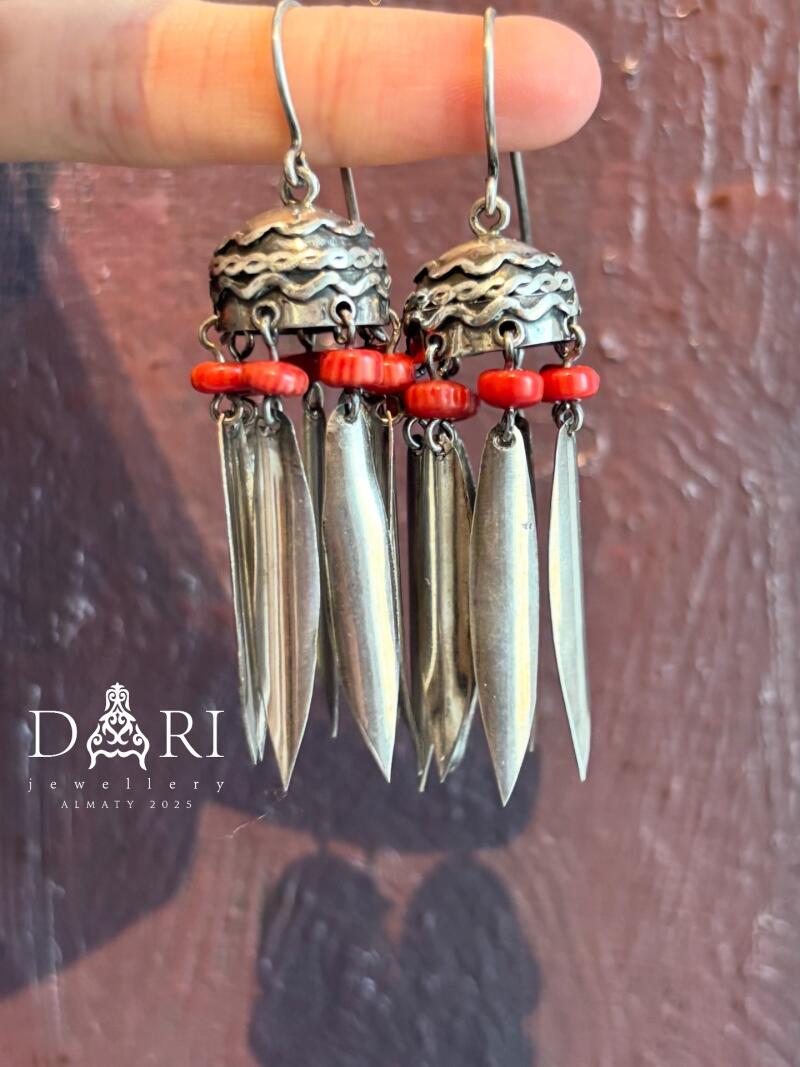 Two silver earrings with red beads and silver pendants are held up against a textured red background; the logo for "DARi jewellery ALMATY 2025" is in the lower-left corner.
