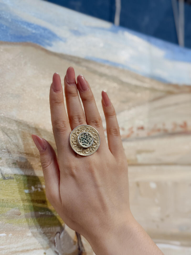 A hand wears a large, ornate silver and gold ring with a circular design.