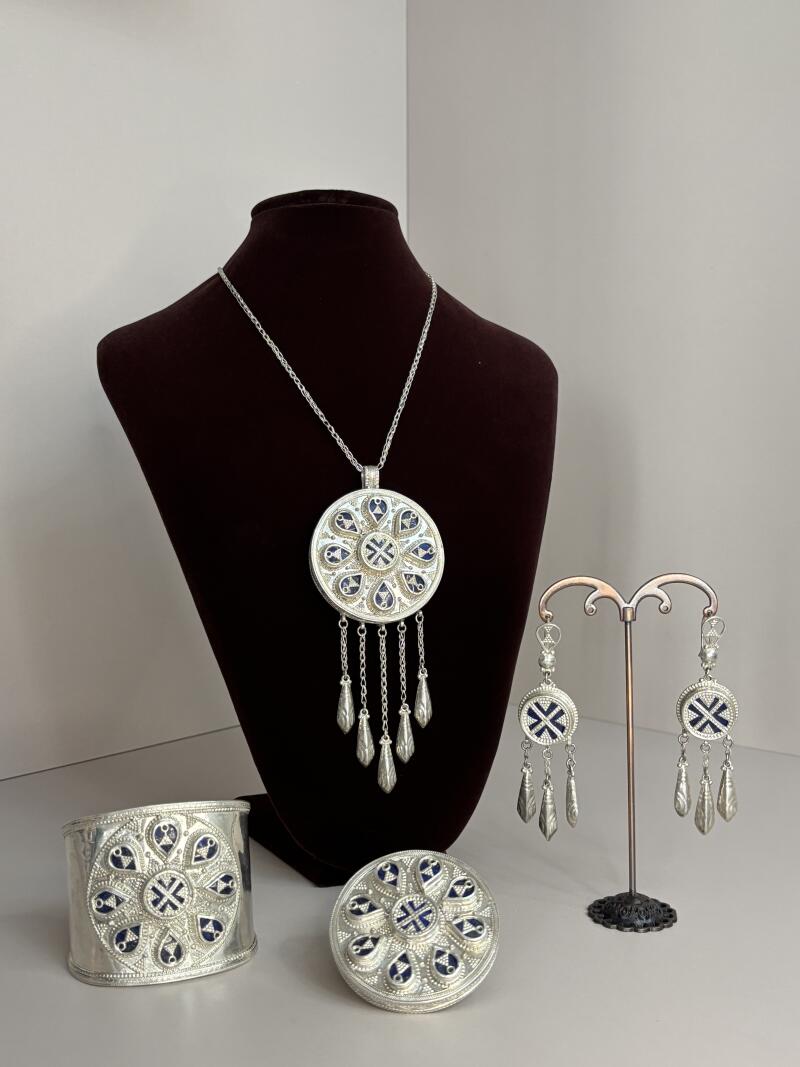 The image shows a set of silver jewelry with blue accents, including a necklace on a brown velvet bust, a bracelet, a ring, and a pair of earrings on a stand.