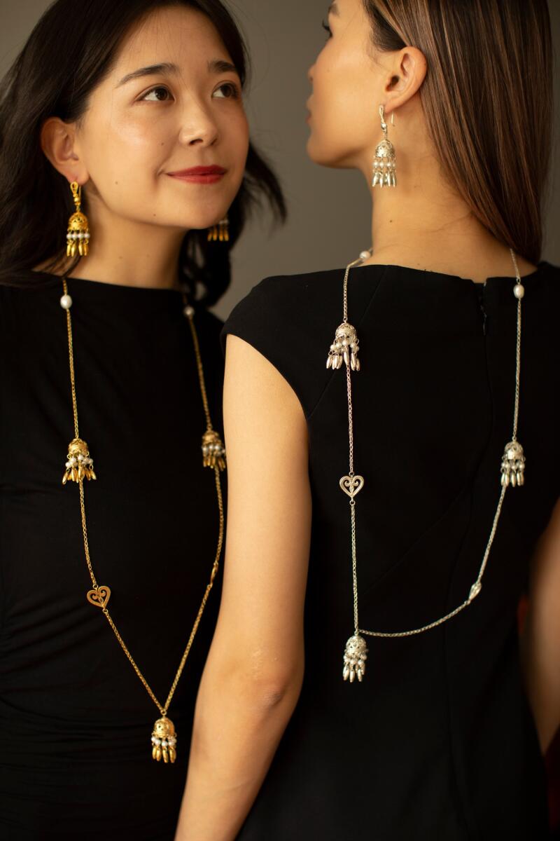 Two women in black dresses are wearing long, ornate necklaces and matching earrings; one necklace is gold and the other is silver.