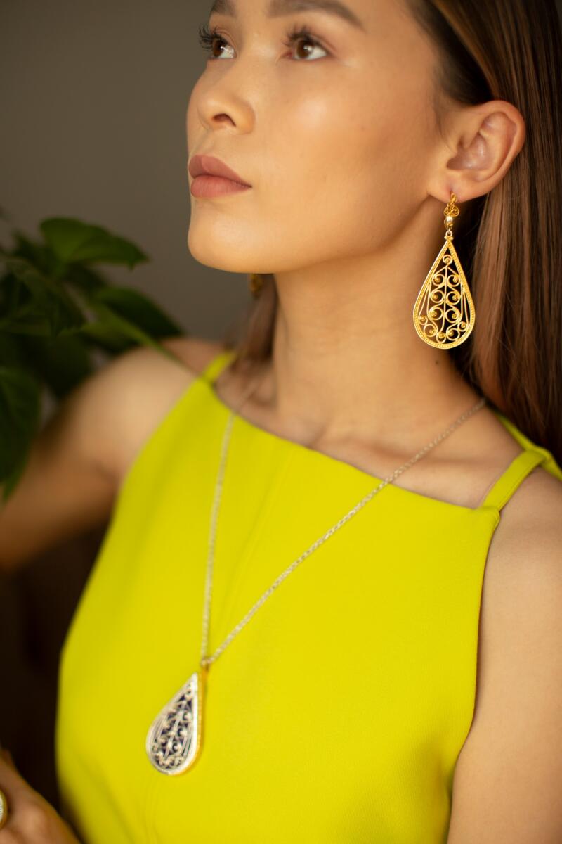 A woman is wearing a yellow dress with gold, filigree teardrop earrings and a teardrop pendant necklace.
