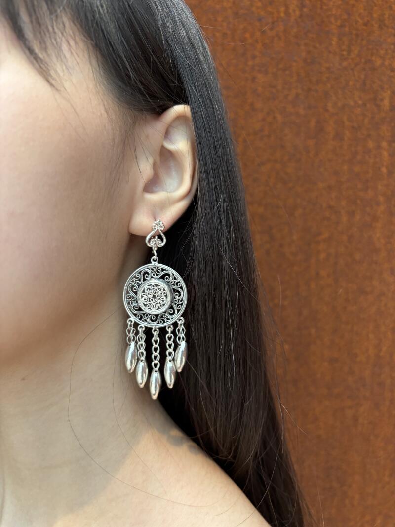 A woman is wearing a silver earring with a circular filigree design and dangling silver teardrop charms.