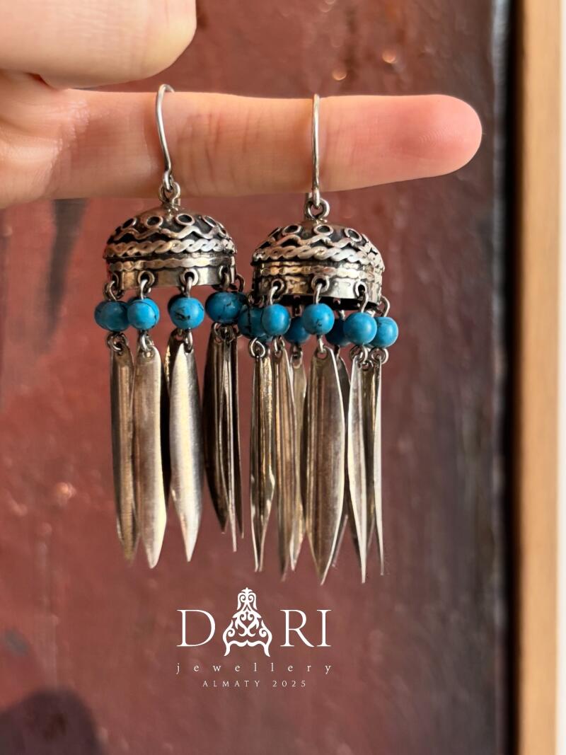 A hand holds up a pair of silver earrings with turquoise beads and long, silver pendants. The earrings are ornate and have a bohemian style. Below, the "DARI jewellery ALMATY 2025" logo is visible.