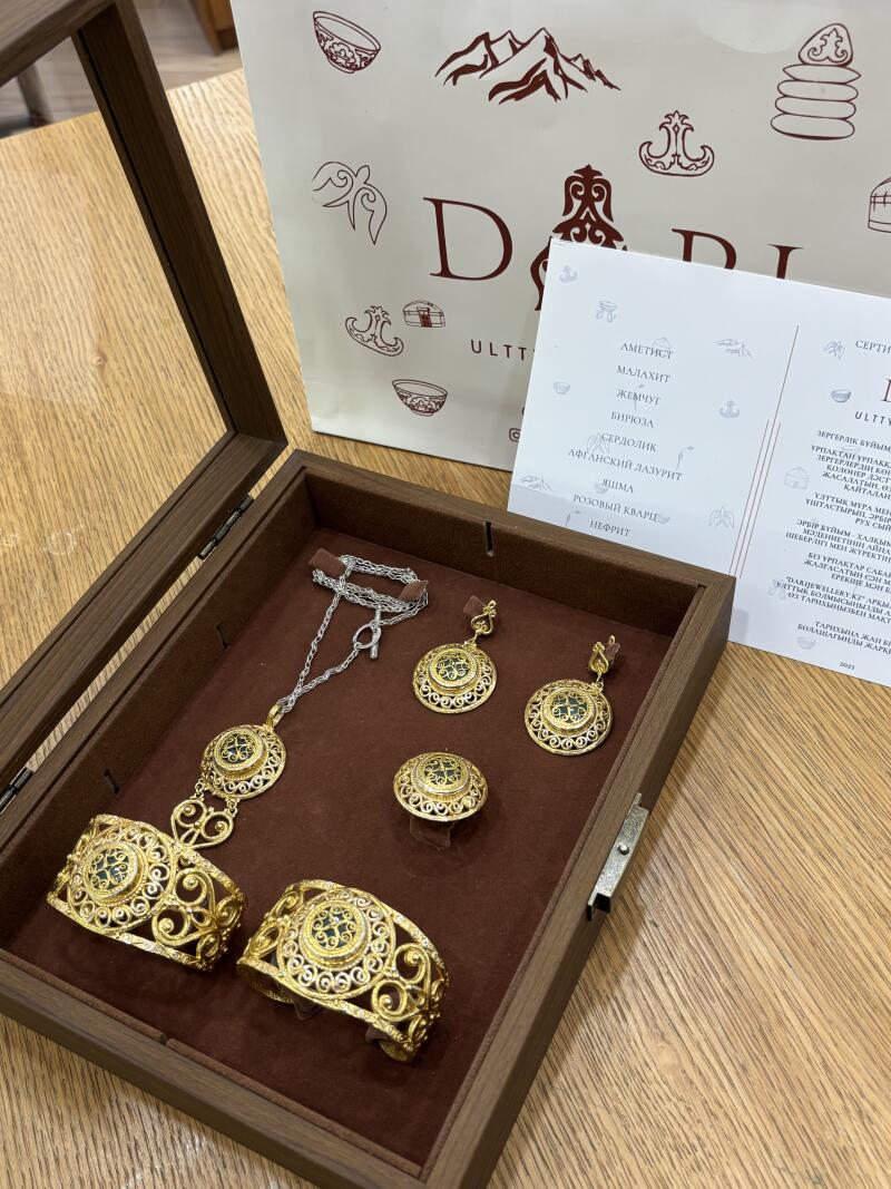 A jewelry set, including a necklace, earrings, a ring, and two bracelets, is displayed in a brown wooden box, with a white bag and a certificate placed behind it. The jewelry is gold with green stones, and the bag features the text 'DI ULTT' along with illustrations.