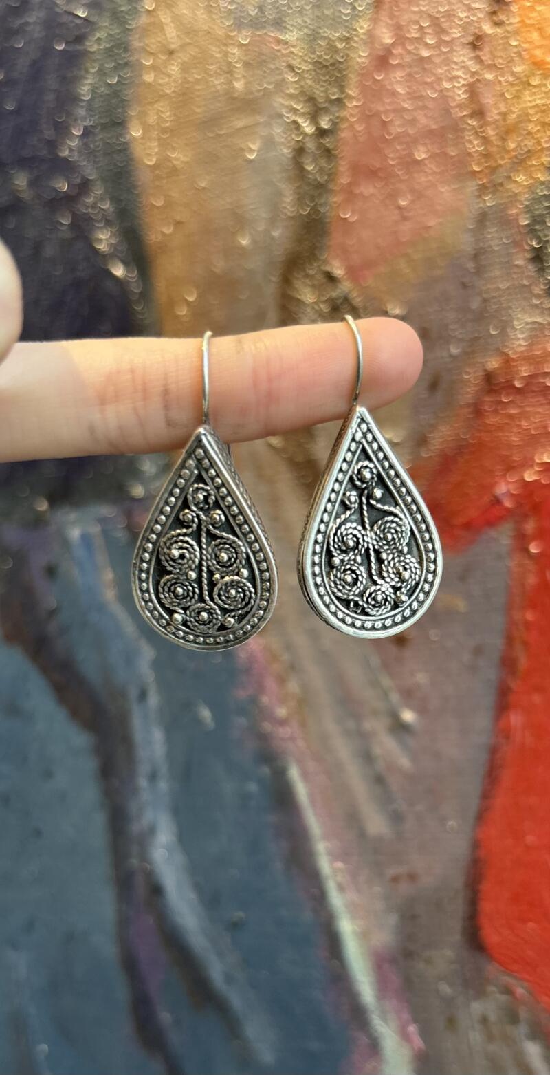 A pair of teardrop-shaped silver earrings with intricate filigree designs is held by a finger.