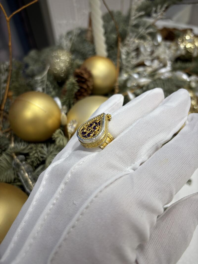 A teardrop-shaped ring with purple stones is worn on a gloved hand, with golden Christmas balls and greenery in the background.