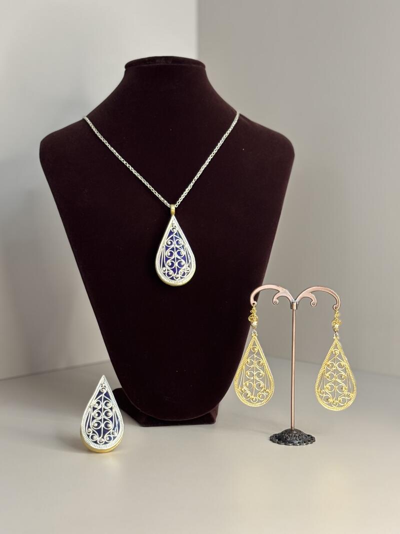 The photo shows a jewelry set consisting of a necklace, a ring, and a pair of earrings, all with a teardrop shape and intricate filigree design with a blue background.