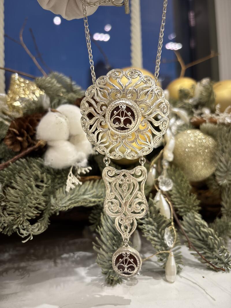 A silver filigree pendant necklace with a maroon center hangs in front of a wintery arrangement of green branches, cotton, and golden ornaments.