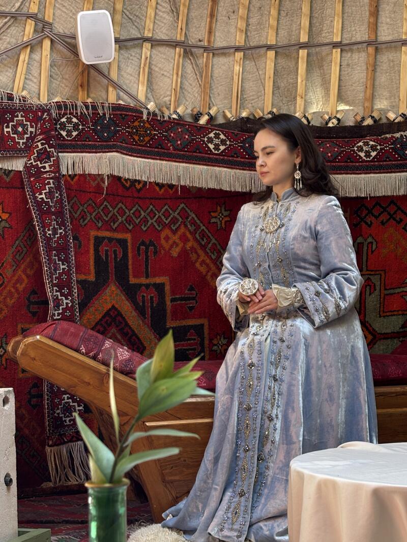 A woman in a traditional Kazakh dress sits inside a yurt, with red patterned carpets hanging on the walls.