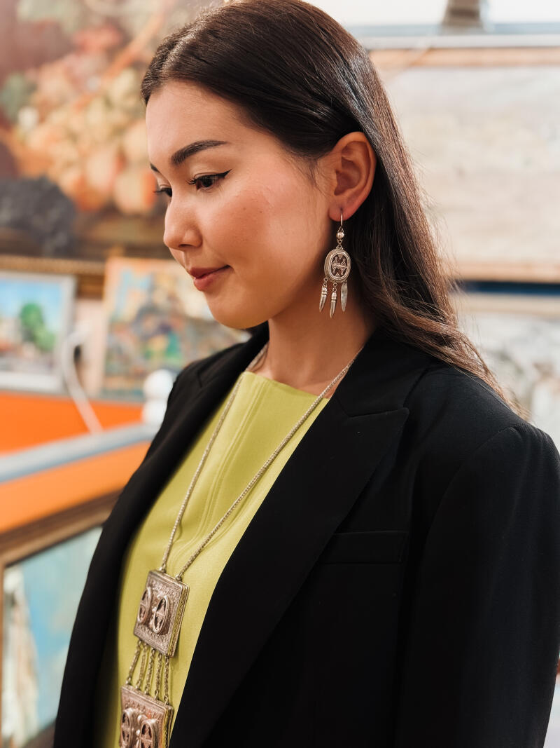 A woman with long dark hair, wearing a green top and a black blazer, is adorned with silver earrings and a necklace with pendant.