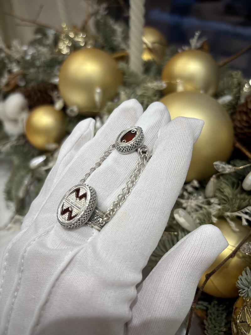 A hand wearing a white glove holds a silver ring connected by a chain to a silver embellishment, set against a backdrop of golden Christmas ornaments and greenery.