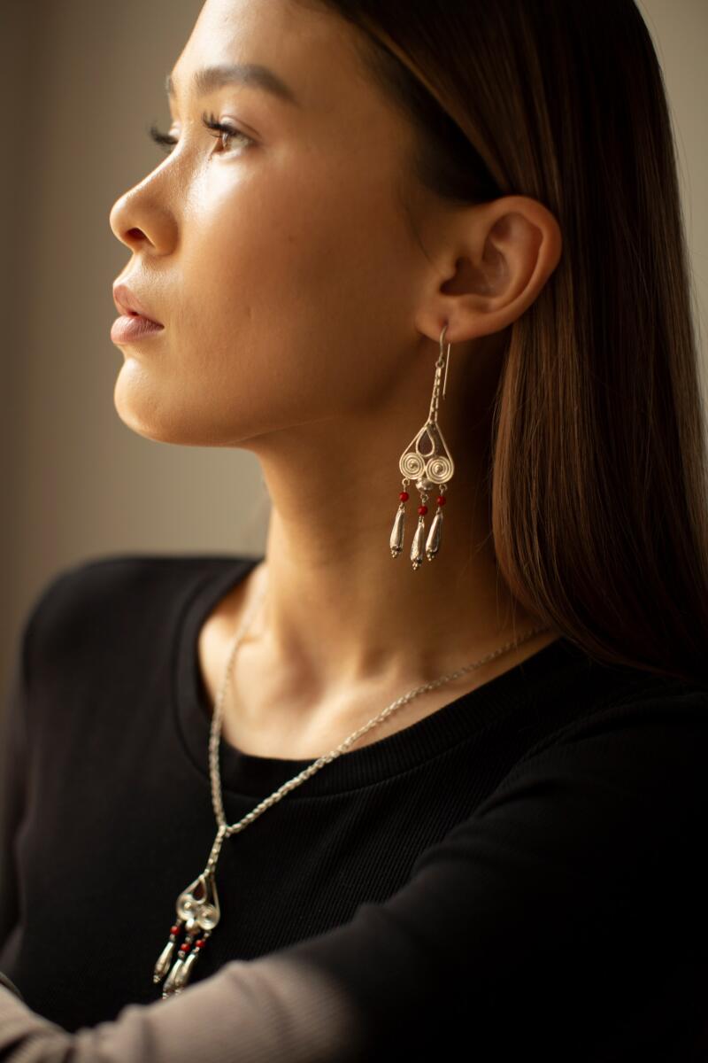 A woman with brown hair wears silver earrings and a matching necklace with red beads, set against a neutral background.