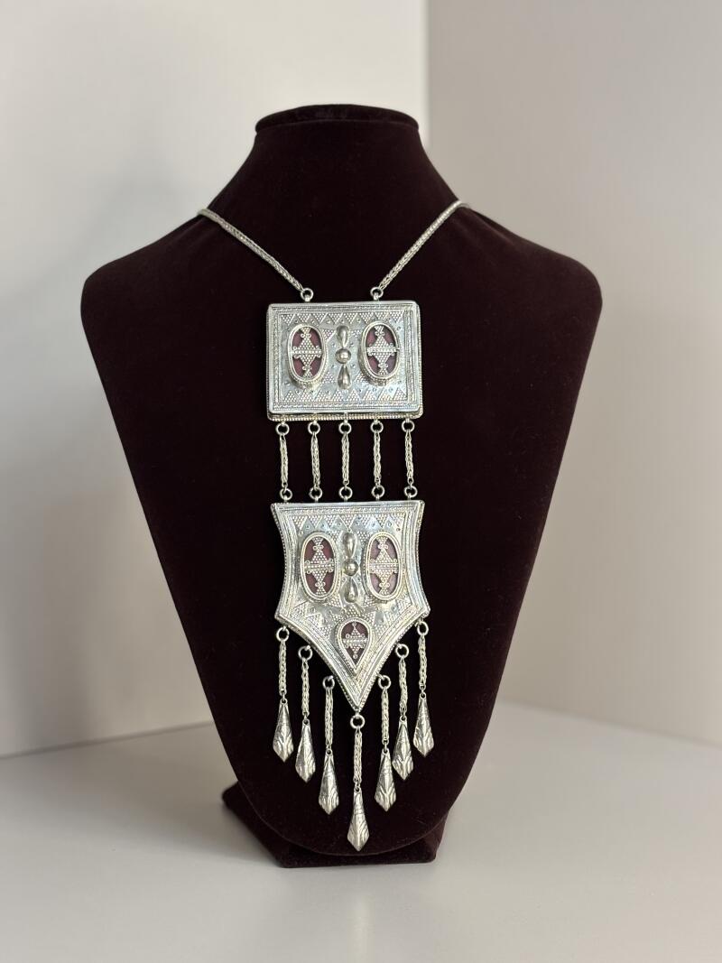 A silver necklace with a rectangular pendant and a shield-shaped pendant hanging from a silver chain on a brown velvet bust.