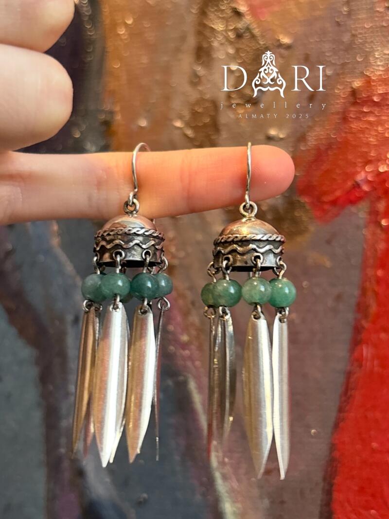 A hand holds two silver earrings with green beads and long, pointed silver tassels, with the logo "DARI jewellery ALMATY 2025" visible in the background.