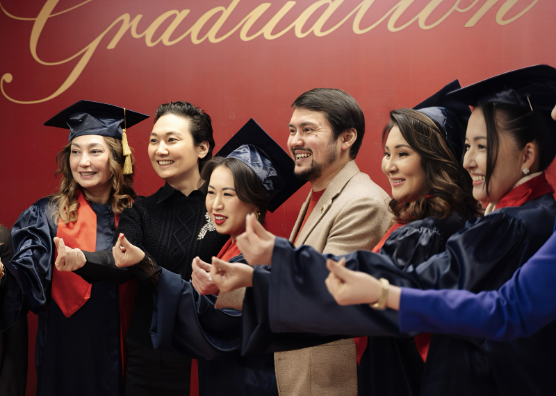 A group of smiling people in graduation gowns and caps pose in front of a red wall with the word "Graduation" in gold script.