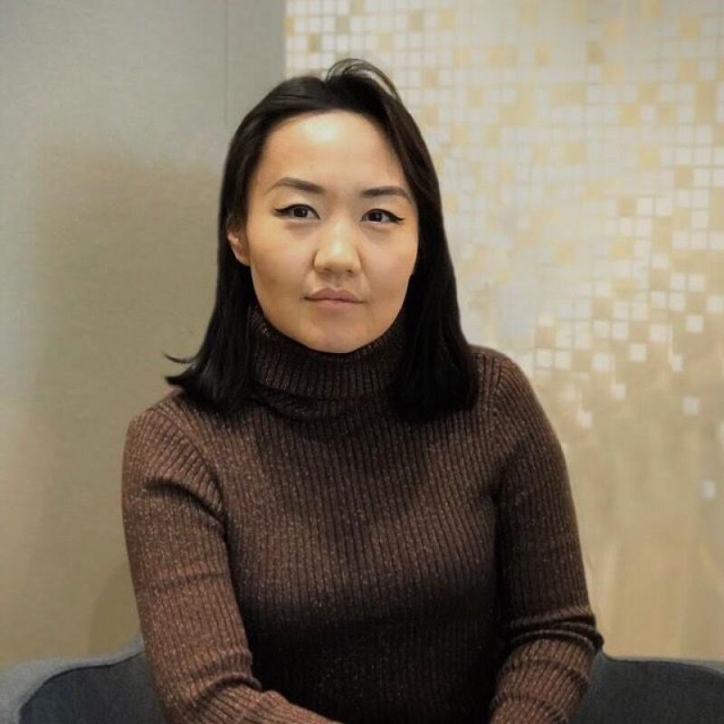 A woman with black hair and eyeliner wearing a brown turtleneck sweater sits in front of a wall with a textured pattern.