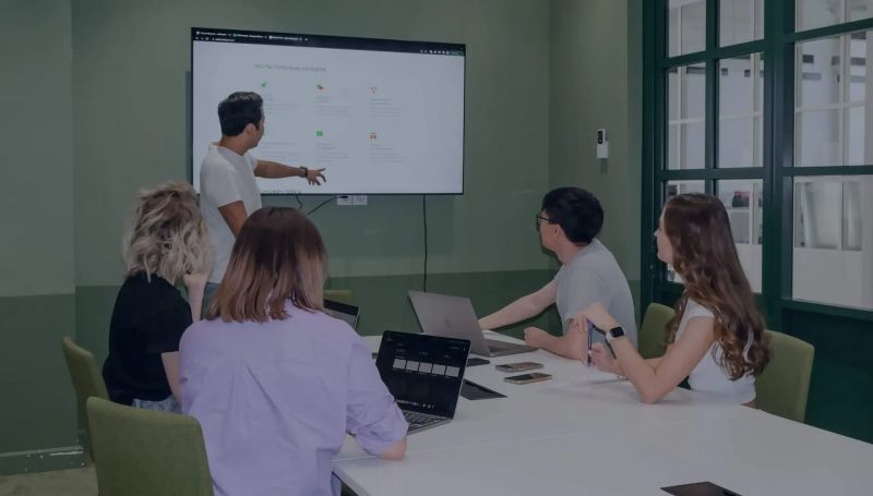 A man points to a screen with the text "ЧТО ТЫ ПОЛУЧИШЬ НА КУРСЕ" while a group of people sit around a table with laptops.