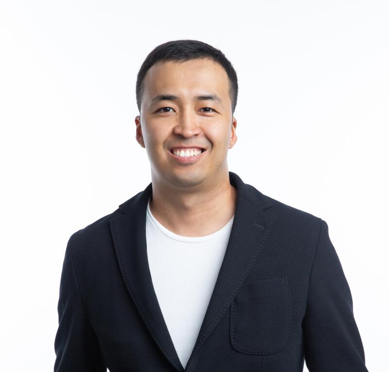 A smiling man with short black hair is wearing a black blazer over a white t-shirt.