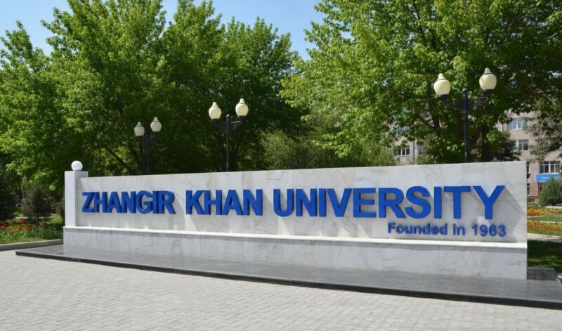 A sign for Zhangir Khan University, founded in 1963, stands in front of green trees and a building.