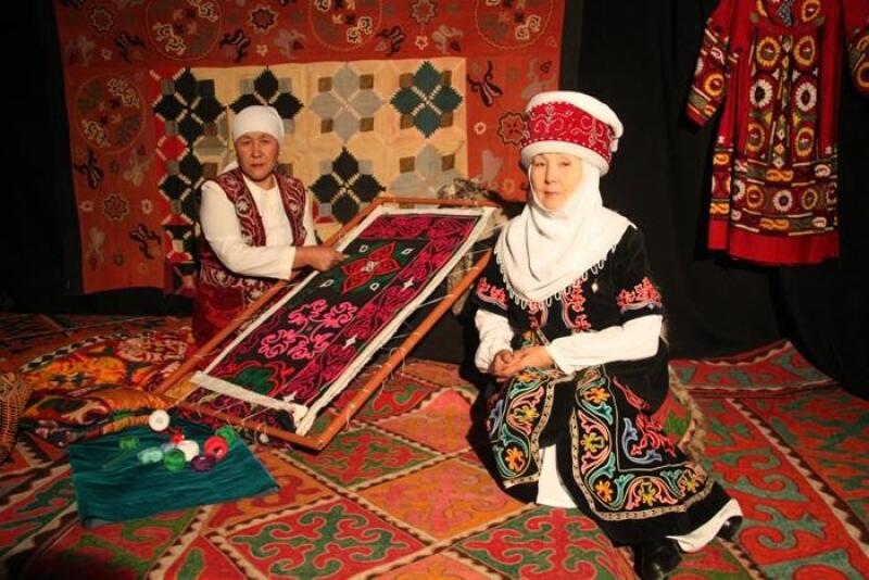 Two women in traditional clothing are on an ornate rug; one is working on a framed textile with colorful threads, and the other is sitting next to her, both against a backdrop of similar textiles.