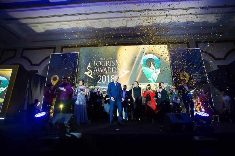A group of people are on stage during the National Tourism Awards 2018 event, with golden confetti falling from above.