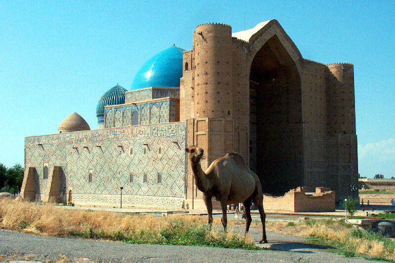 A camel stands in front of the Mausoleum of Khoja Ahmed Yasawi, which has a large arched entrance and blue domes.