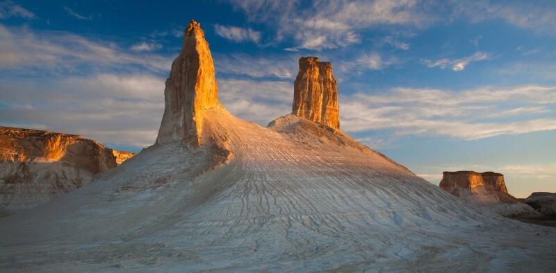 The landscape features tall, jagged rock formations under a partly cloudy blue sky, with sunlight casting shadows on the sandy terrain.