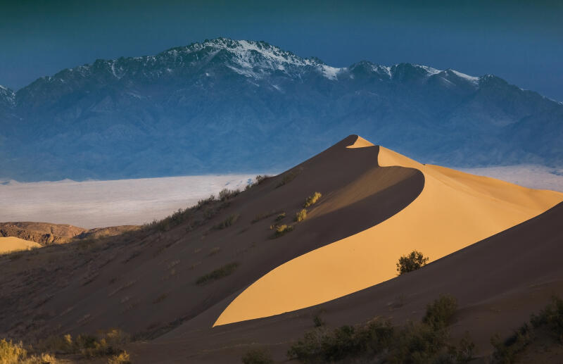 The landscape features a large sand dune in the foreground, with sparse vegetation, against a backdrop of distant, snow-capped mountains under a twilight sky.
