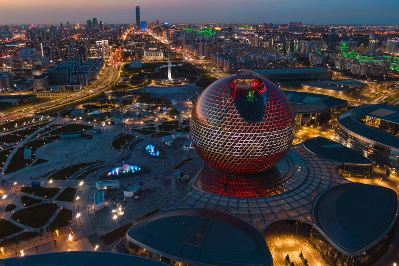 An aerial view captures the illuminated cityscape at dusk, dominated by a large, spherical building adorned with a grid pattern, set amidst a sprawling urban park and modern architecture.