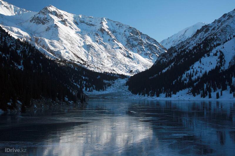 The landscape shows a frozen lake surrounded by snow-covered mountains and dark green trees, under a clear blue sky. The text "iDrive.kz" is visible at the bottom left.