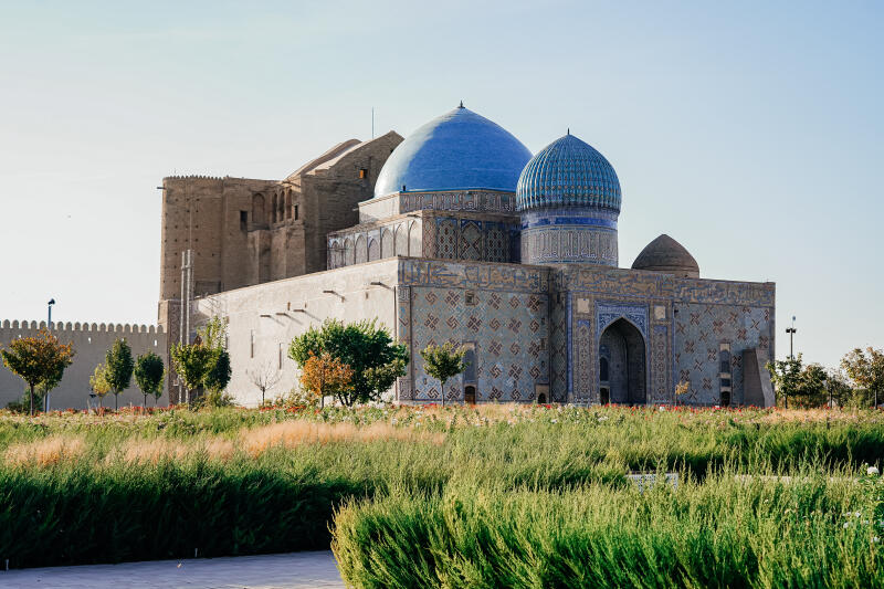 An exterior view of a large, ornate building with blue domes and intricate tile work, surrounded by green bushes and small trees under a clear sky.