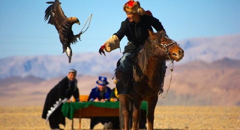 A man in traditional clothing rides a horse, releasing an eagle into the air, while other people sit at a table in the background.