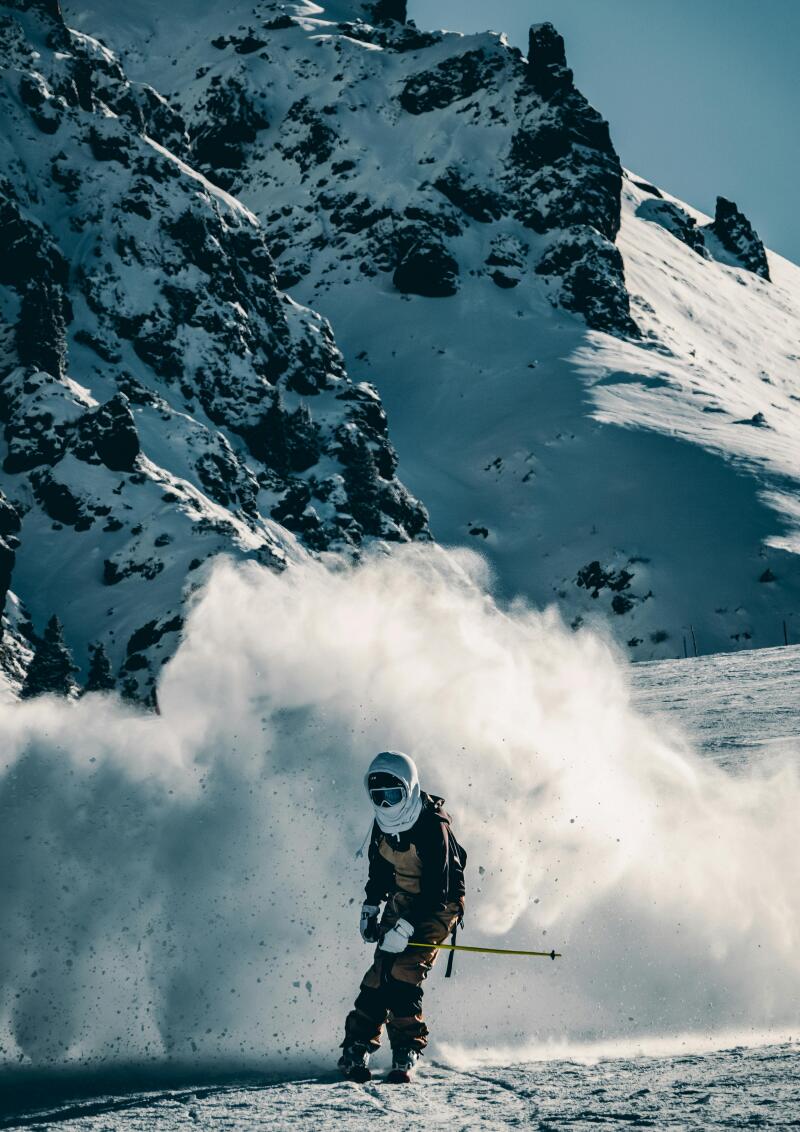 A skier in full gear is skiing down a snow-covered mountain, kicking up a cloud of snow, with a large, rocky mountain in the background.