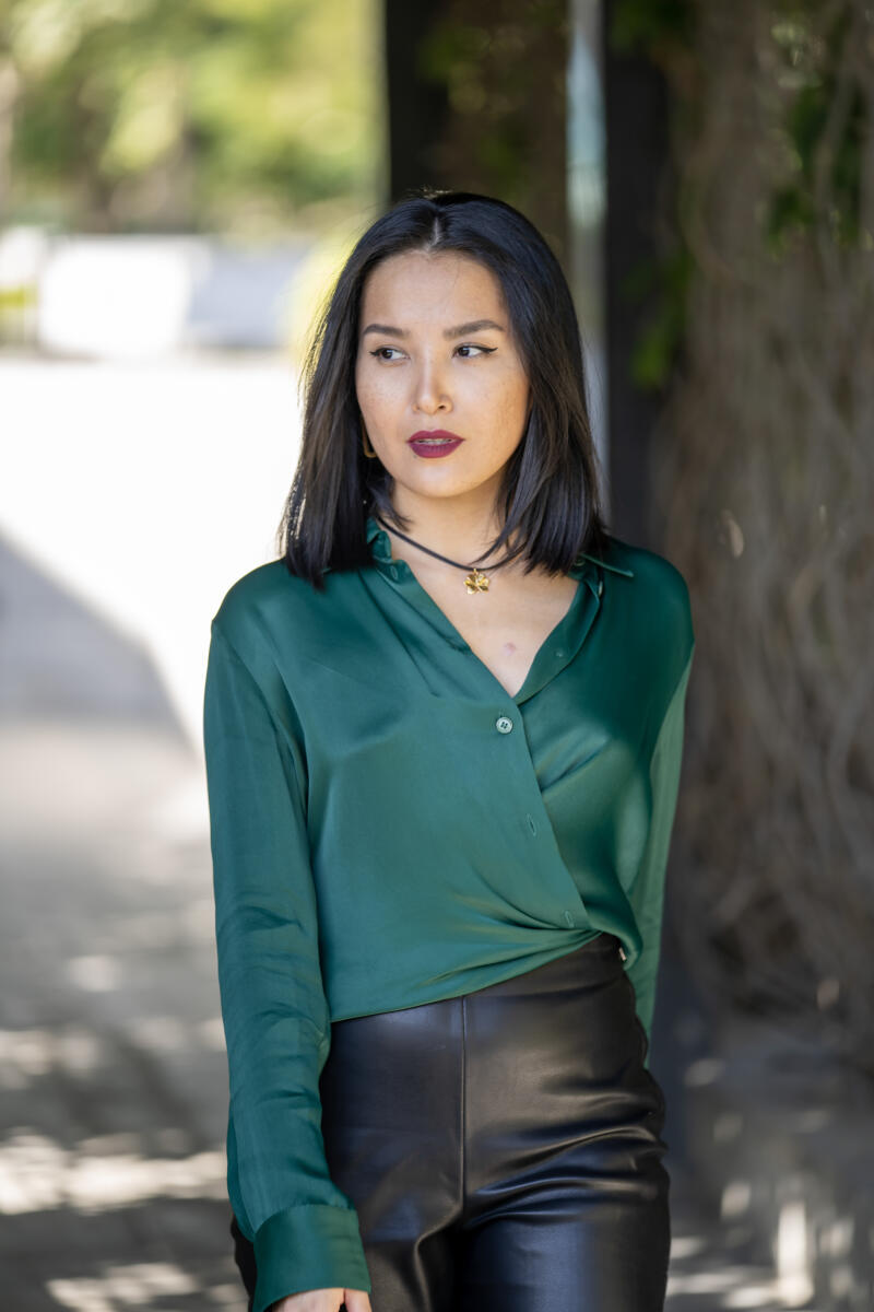 A woman with dark hair and dark red lipstick is wearing a green silk shirt and black leather pants.