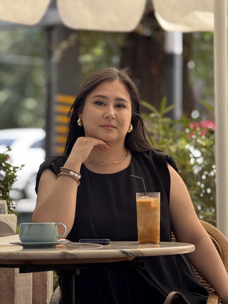 A woman is sitting at a table with a cup of coffee and a glass of iced coffee, wearing a black dress and gold jewelry.