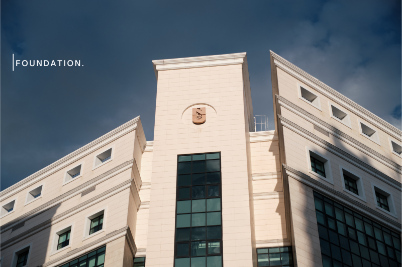 A low-angle shot of a cream-colored building with large windows under a cloudy sky, with the word "FOUNDATION." on the left.