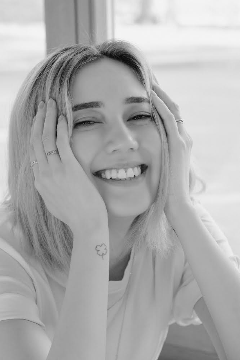 A black and white portrait of a woman smiling with her hands on her face, showing rings and a clover tattoo on her wrist.
