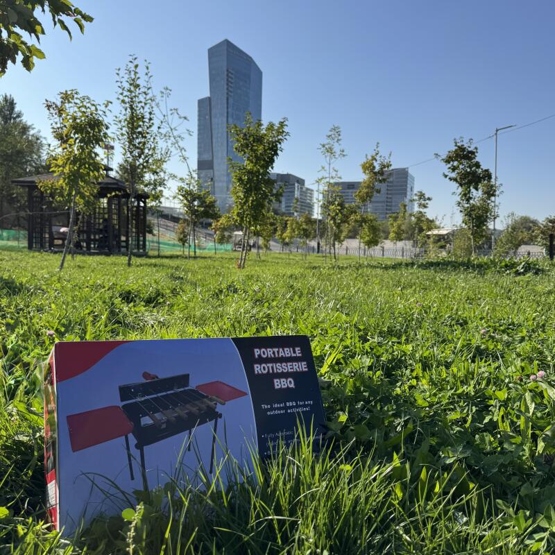 In a grassy field, a box for a 'Portable Rotisserie BBQ' is prominently displayed, with the text 'The ideal BBQ for any outdoor activities!' visible on the packaging; in the background are trees, a gazebo, and city buildings under a clear sky.