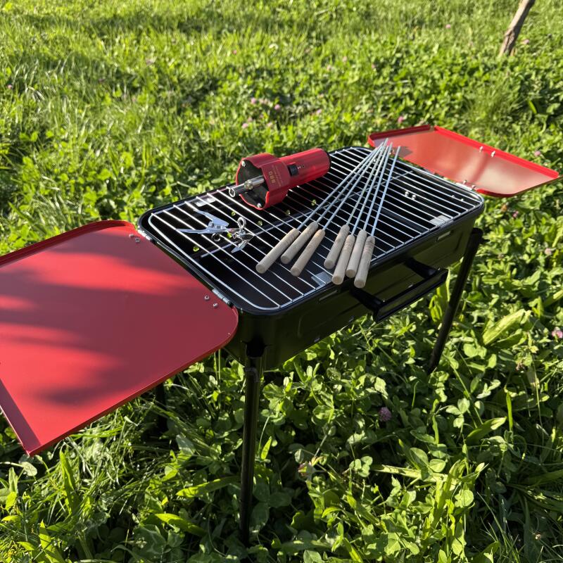 A black charcoal grill stands on green grass, featuring red side tables, skewers with wooden handles, a red electric motor, and metal tongs.