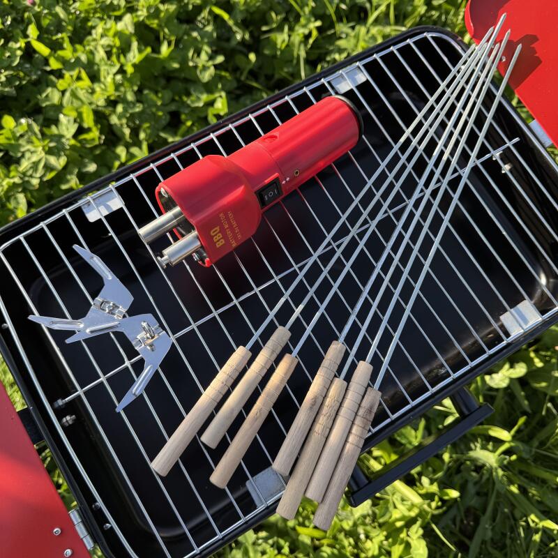 The image shows a black barbecue grill with a red rotisserie motor labeled 'BBQ BATTERY MOTOR DC 1.5V BATTERY', metal skewers, wooden-handled skewers, and a metal clamp on top of green grass.