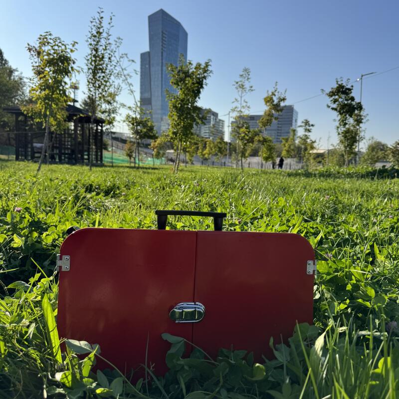 A closed red portable grill sits in a grassy field, with trees and a building in the background under a blue sky.