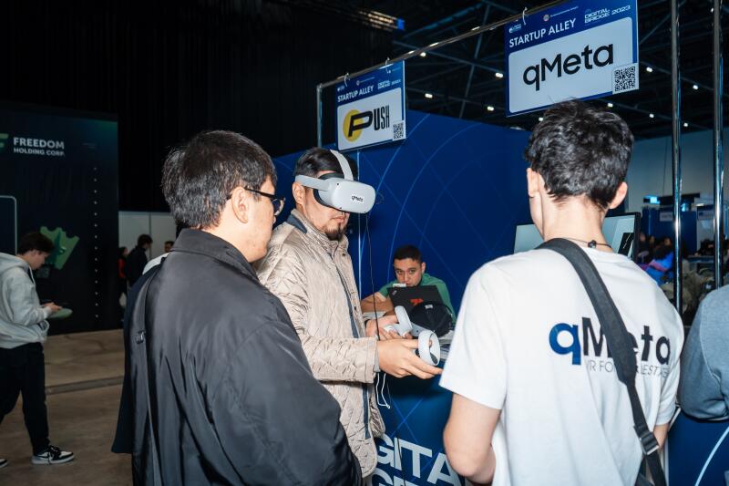 At a tech event, a man wearing a VR headset holds controllers while standing near "Startup Alley" booths for "qMeta" and "PUSH", with the text "FREEDOM HOLDING CORP" visible in the background.